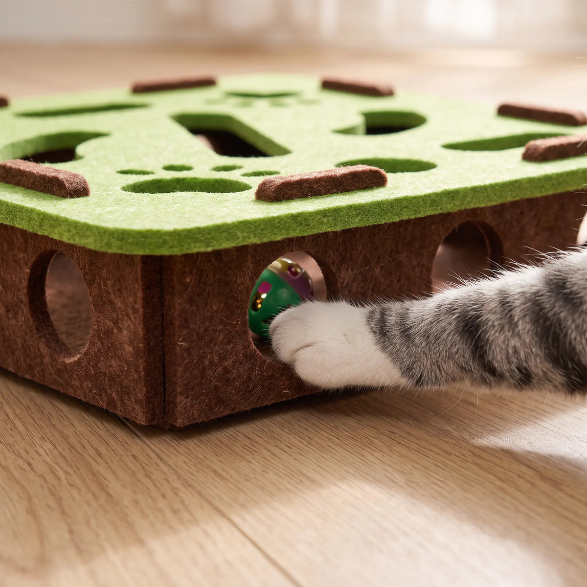 Patte de chat engagée dans un trou latéral du caisson en feutre brun pour atteindre une balle à grelot